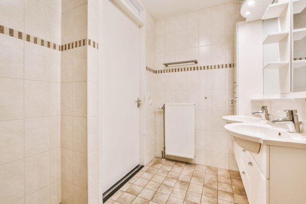 Outdated bathroom with double sinks, vanity cabinets, tile flooring, and older fixtures before renovation.